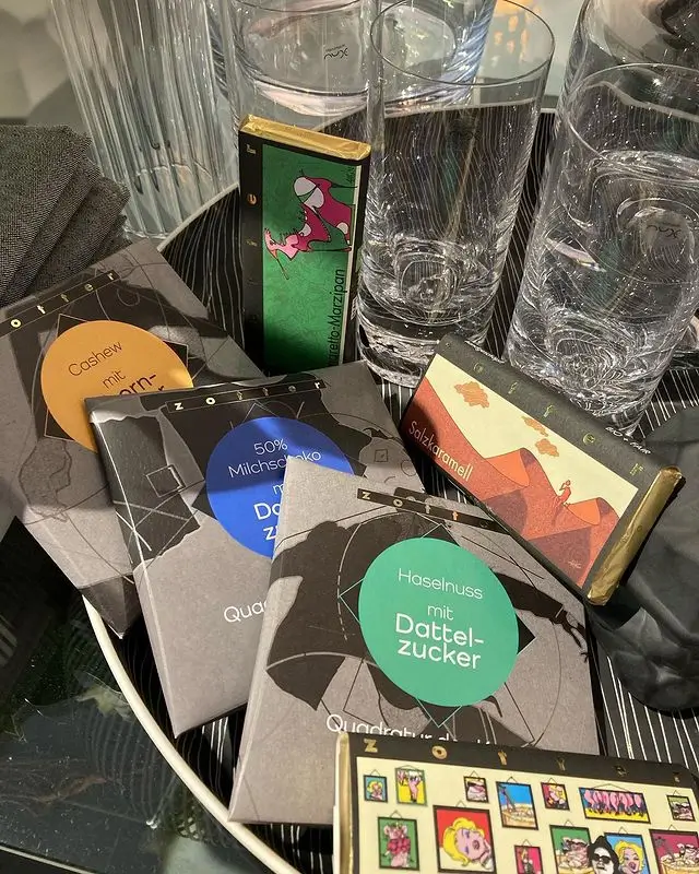 Mehrere bunte Tafeln Schokolade mit besonderen Zutaten liegen auf einem schwarzen Tablett neben zwei Gläsern Wasser und einem Stapel dunkler Servietten