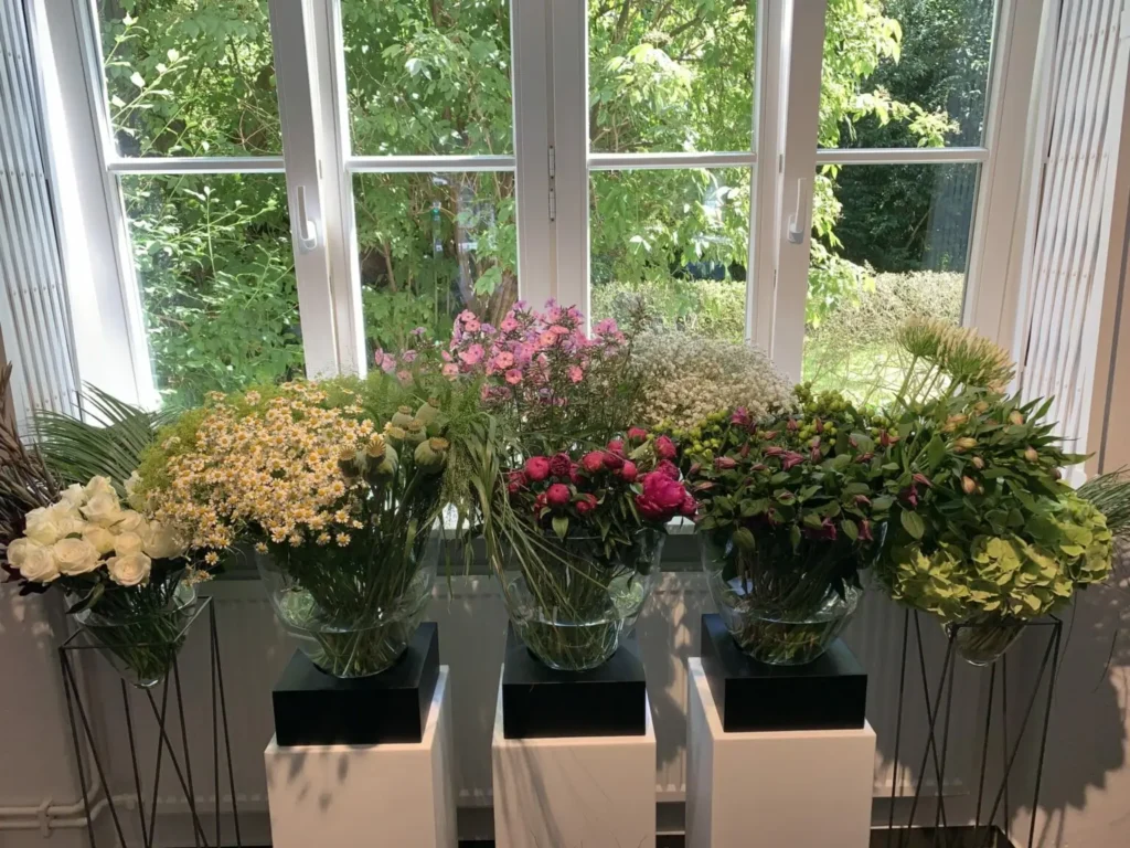Fuenf Glasvasen mit unterschiedlichen bunten Blumen stehen vor einem Fenster mit Gruenblick